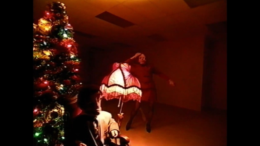 A dimly lit room featuring a Christmas tree adorned with colorful lights on the left, while a woman in a red dress dances near a vintage lamp on the right, with a mannequin seated in the foreground.