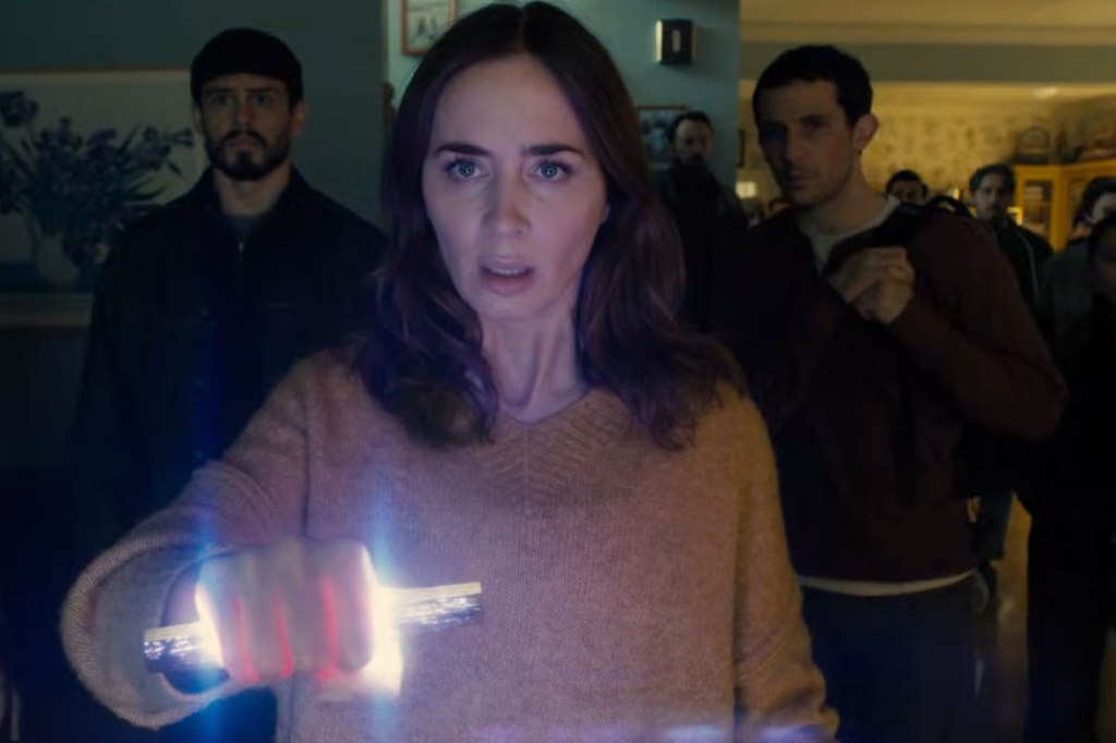 A woman in a brown sweater holds a glowing object in her hand, looking concerned. Two men stand behind her, watching intently.