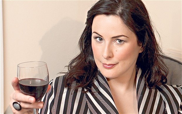 A woman in a striped robe holding a glass of red wine, looking confidently at the camera.