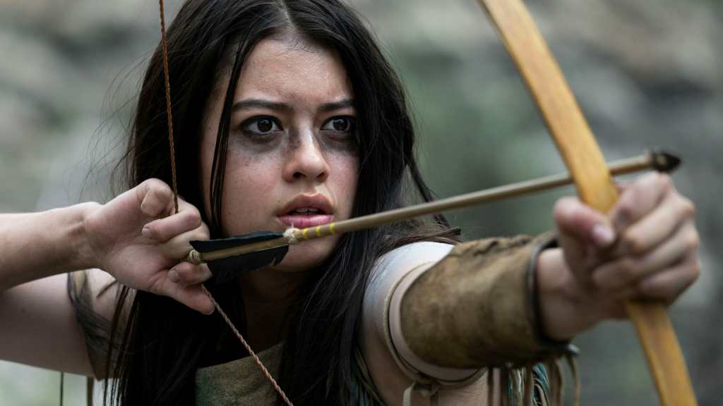 A young woman with long dark hair and dirt smudges on her face is aiming a bow and arrow with a determined expression.