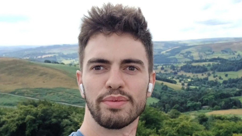 A young man with short hair and earbuds stands in front of a panoramic landscape featuring rolling hills and lush greenery.
