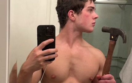 A young man taking a mirror selfie while holding a hammer, shirtless and looking to the side.