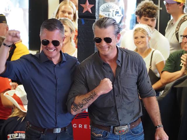 Two men in sunglasses smiling and posing with raised fists, surrounded by a crowd in a lively setting.