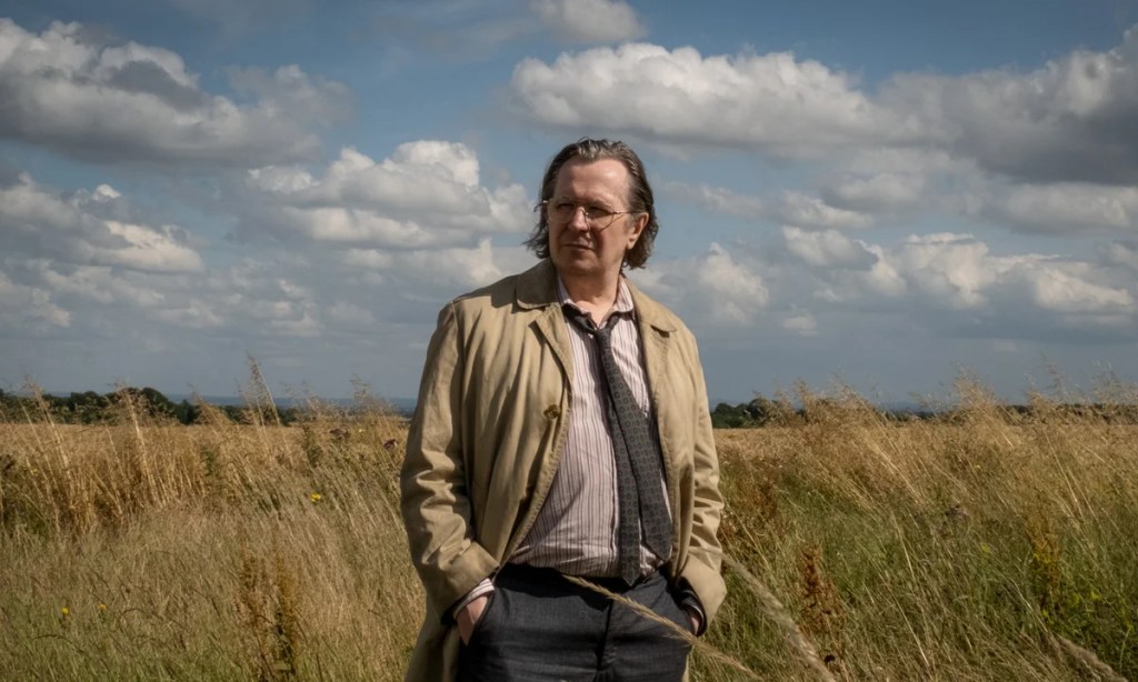 A man standing in a field of tall grass under a partly cloudy sky, wearing a light-colored coat and a striped shirt with a tie.