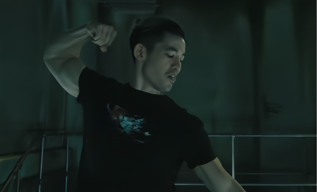 A man flexing his arm muscles while wearing a black t-shirt, set in a dimly lit room with industrial elements.