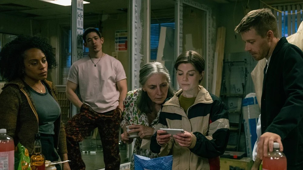 Five individuals gathered in a room, focusing on a smartphone screen. Two women are examining the phone closely, while a man stands nearby. The setting is cluttered, with various drinks and food items on a table.