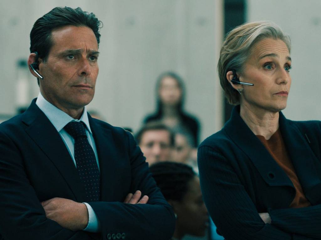 Two business professionals with earpieces standing with arms crossed, looking serious in a corporate setting.