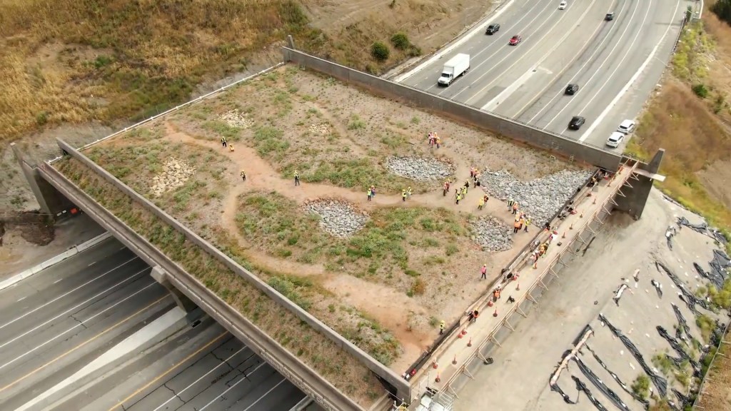 Aerial view of a green space on top of a highway overpass, with construction workers in safety vests walking on a dirt path. Surrounding areas have rocky terrains and vegetation.